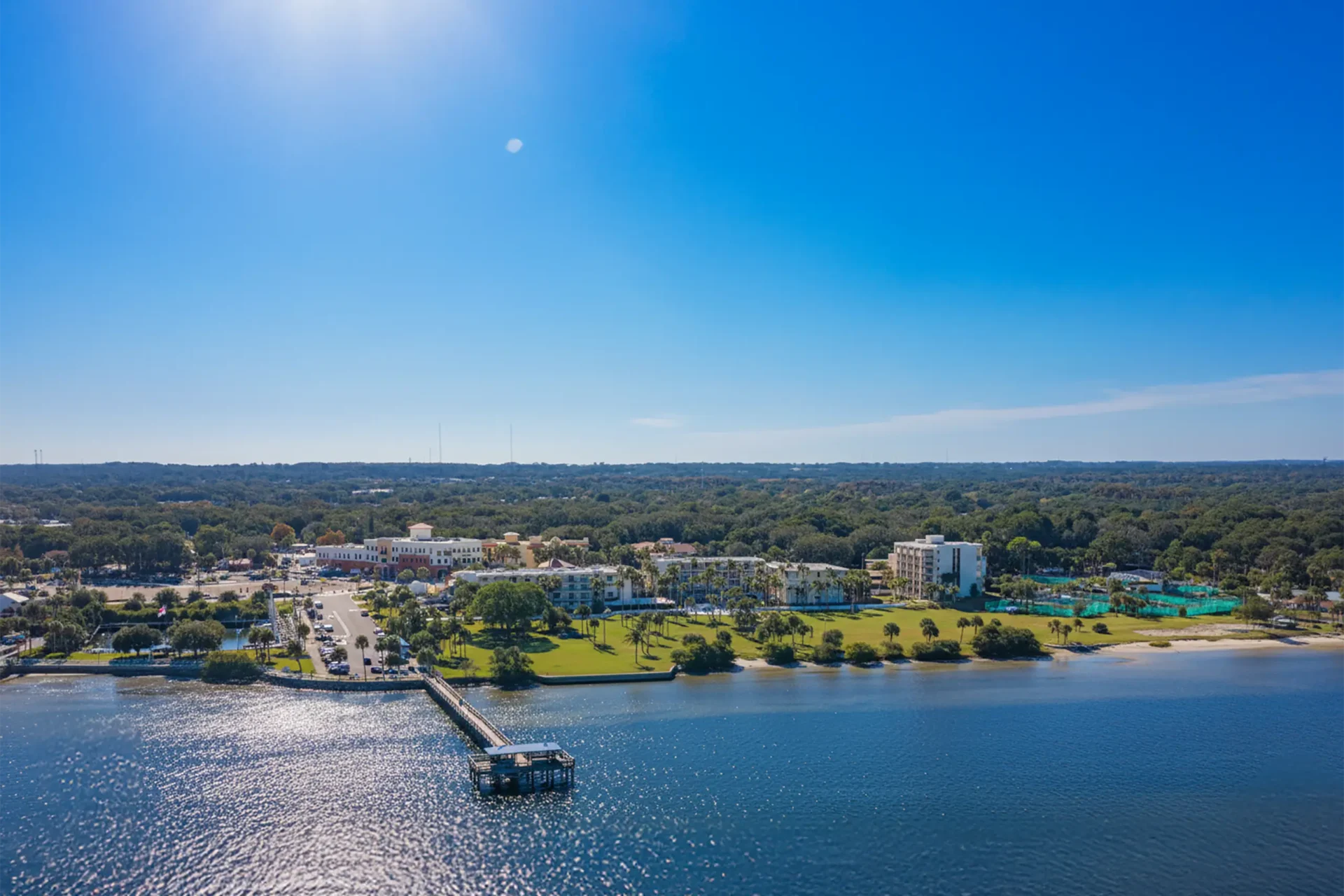 Aerial view of a waterfront with a long pier extending into shimmering blue water, sun glare on the surface and a green park along the shore.