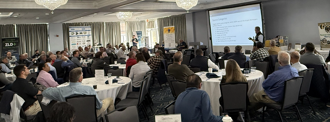 Audience seated at round tables in a conference room, presenter at the front delivering slides projected on a screen.