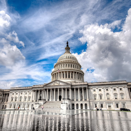 U.S. Capitol building in Washington, D.C., with dome and marble steps, reflected in a wet foreground pool under a blue, cloudy sky.